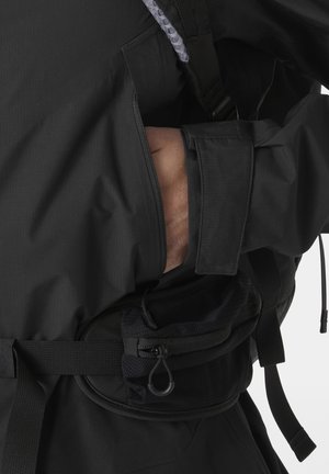 Hand in de zijzak van een zwart jack met gespsluiting aan de mouw en een zwarte heuptas met ritsvakken.