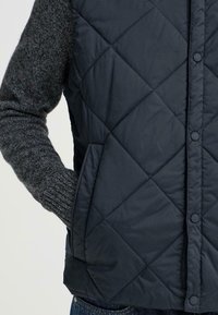 Fodrad marinblå väst med ett slätt yttermaterial och grå strumpärmar, med tryckknappar och sidofickor för ökad funktionalitet.