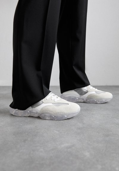 White suede sneakers with a transparent sole and grey accents, paired with black trousers, on a concrete surface.