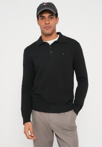 Mustast kudumist pullover kraega ja kolme nupuga, millel on soonikkoes mansetid ja alumine äär. Vasakul rinnal väike logo. Kandmiseks on sobitatud hallide pükstega.