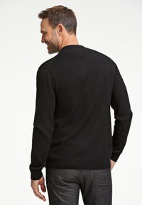 Mustat neulotut neuleet, joissa on ribbineuletta kauluksessa ja hihoissa. Pehmeä rakenne, istuva muotoilu, yhdistetty harmaisiin teksturoituihin housuihin, näkyy takaa.