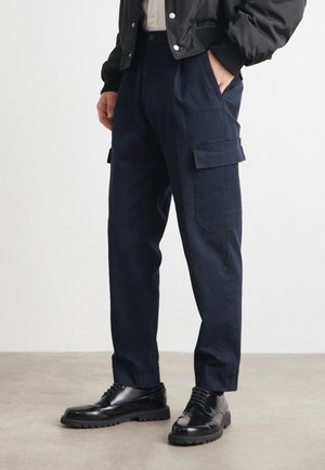 Pantaloni cargo bleumarin cu două buzunare laterale, confecționați dintr-un material rezistent. Asortați cu cizme negre cu șireturi, având o talpă robustă.
