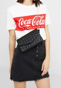 Vävd svart axelremsväska med klaffstängning, silverfärgad kedjerem och slät textur, som bärs över en vit och röd t-shirt med Coca-Cola-logotyp.