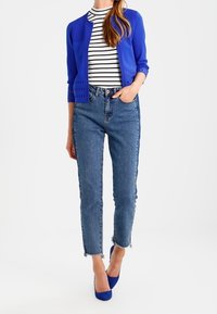 Kvinna i en kortad blå cardigan över en svartvit randig topp, högmidjade blå jeans med fransade sömmar och blå klackar, som står med en hand i fickan.