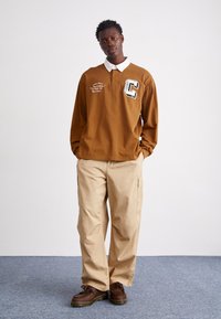 Brun långärmad polo med vit krage och logotyp, tillsammans med beiga cargobyxor. Modellen bär bruna snörskor.
