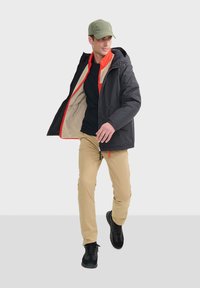 Schwarze und rote Zip-Jacke mit Kapuze, beiger Innenausstattung, kombiniert mit einem schwarzen T-Shirt und beigen Hosen, abgerundet mit schwarzen Sportschuhen.