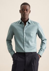 Chemise à manches longues bleu-vert clair avec un devant boutonné et un col classique. Associée à un pantalon sombre, avec une texture de tissu lisse.