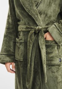 Accappatoio in morbido verde oliva con cintura in vita, due tasche frontali e una texture soffice. Presenta un colletto sciallato e maniche larghe.