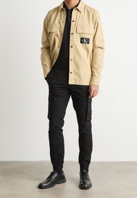 Beżowa koszula na guziki z dwoma przednimi kieszeniami i czarną naszywką z logo, zestawiona z czarnymi spodniami cargo z bocznymi kieszeniami i elastycznymi mankietami. Czarne buty.