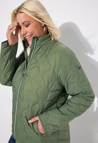 Giubbotto trapuntato verde con colletto alto e chiusura a zip. Presenta tasche, un motivo esagonale e una toppa sulla manica sinistra.