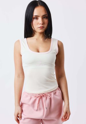 Jeune femme aux longs cheveux foncés, portant un haut blanc sans manches et un pantalon rose clair à cordon, debout devant un fond uni.