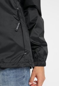 Sort jakkeærme med elastisk manchet vist med hånd ved siden af hoften, med en lynlåslomme mærket "Pack Pocket" på lyseblå denim jeans baggrund.