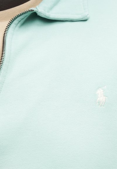 Svetlo modri pulover iz pletenine s podaljšanim zadrgo v srebrni barvi in majhnim belim vezenim logotipom konja in jahača na prsih.