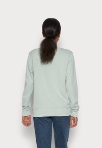 Persona con cabello oscuro en una coleta baja, vistiendo un suéter verde claro y jeans azules, de pie de espaldas contra un fondo simple.