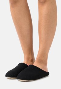 Black knitted slippers with a rounded toe and textured exterior. The interior features a pink accent. Flexible sole for comfort.