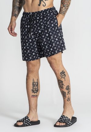 Mężczyzna ubrany w czarne, wzorzyste szorty i dopasowane klapki, prezentujący liczne tatuaże na ramionach i nogach, stojący na tle jednolitego tła.