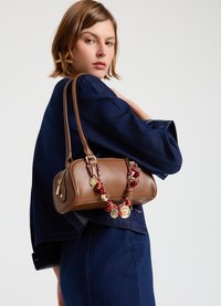 Braune Lederhandtasche mit abgerundeter Form, kurzen Griffen und dekorativer Kette mit roten Perlen und goldenen Akzenten. Getragen über einer Jeansjacke.