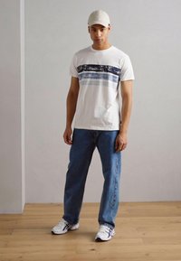 Weißes T-Shirt mit blauen horizontalen Mustern, kombiniert mit lockeren blauen Jeans und weißen Sneakers. Die Person trägt außerdem eine hellfarbige Kappe.