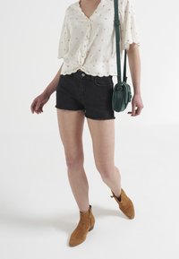 Black denim shorts with frayed hem, paired with a white blouse featuring scalloped edges and gold accents. Worn with tan ankle boots and a green bag.