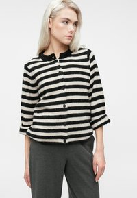 Blondynka w czarno-białym paskowanym swetrze z guzikami i podwiniętymi rękawami oraz ciemnoszarymi spodniami, patrząca w prawo na białym tle.