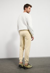 Hellgrauer Sweatshirt, beigefarbene Jogginghosen mit elastischen Bündchen, Taschen detail und schwarz-grüne Sportschuhe mit reflektierenden Akzenten.
