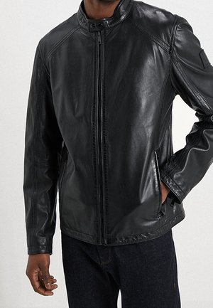 Homme portant une veste en cuir noire zippée avec col montant, main dans la poche, vêtu d'un jean foncé.