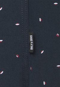 Tessuto blu navy con piccoli motivi astratti rossi e bianchi. Presenta un'etichetta nera con "ONLY SONS" in bianco, posizionata vicino alla cucitura.