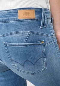 Primer plano del bolsillo trasero y la cintura de unos vaqueros de mezclilla azul claro con una etiqueta de marca de cuero marrón y diseño de bolsillo cosido.