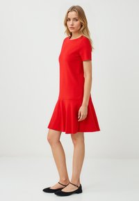 Une jeune femme pose dans une robe évasée rouge vif et des ballerines noires sur un fond blanc minimaliste. Ses cheveux sont ondulés et lâches.