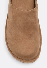 Suede natikače svijetlosmeđe boje. Zaobljen vrh s glatkom teksturom, suptilni detalj šivanja duž gornjeg dijela i podstavljeni đon.