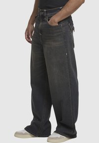 Luźne, ciemne dżinsy o efekcie wyblaknięcia, z głębokimi kieszeniami i zamkiem błyskawicznym, w zestawie z białymi i szarymi sneakerami.