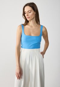 Top de tirantes azul acanalado con escote cuadrado, combinado con pantalones blancos de cintura alta y plisados. Textura suave, forma ajustada y sin patrones visibles.