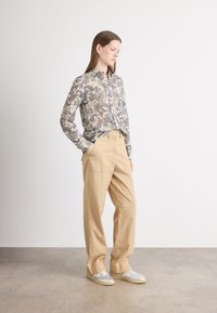 Camisa de manga longa abotoada com um padrão paisley em bege, azul e castanho. Calças bege de corte solto. Ténis cinzentos com atacadores brancos.