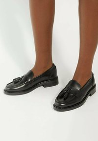 Black leather loafers with tassel accents, rounded toe, and low block heel. Smooth texture with stitching details along the edges.
