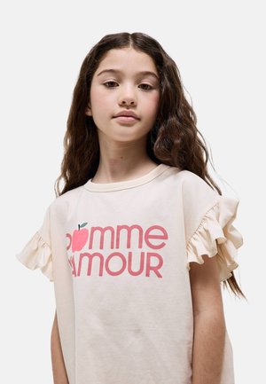Jeune fille aux longs cheveux ondulés portant un t-shirt crème à manches volantées avec un texte rose "pomme amour" accompagné d'un graphique de pomme.