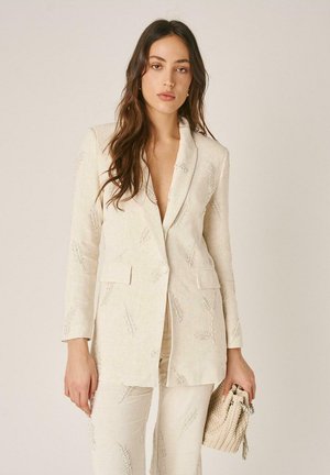 Mujer con blazer y pantalones texturizados en color crema con sutiles patrones bordados de hojas, sosteniendo un bolso de mano tejido en color crema.