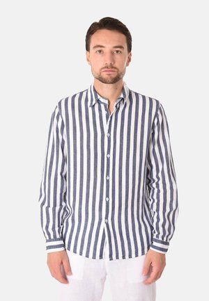 Uomo che indossa una camicia a maniche lunghe abbottonata con righe verticali blu navy e bianche e pantaloni bianchi, in piedi davanti a uno sfondo uniforme.