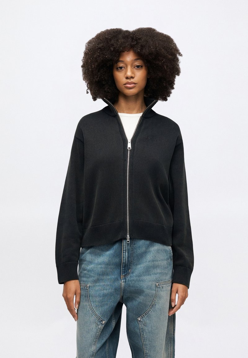 Black zip-up sweater with ribbed hem and cuffs, fitted design, made from a smooth texture, paired with light blue denim jeans.