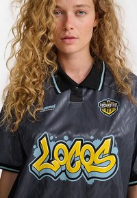 Maillot style polo noir avec un motif géométrique, comportant un graphique jaune et bleu "Locos" bien visible ainsi qu'un logo sur la poitrine.