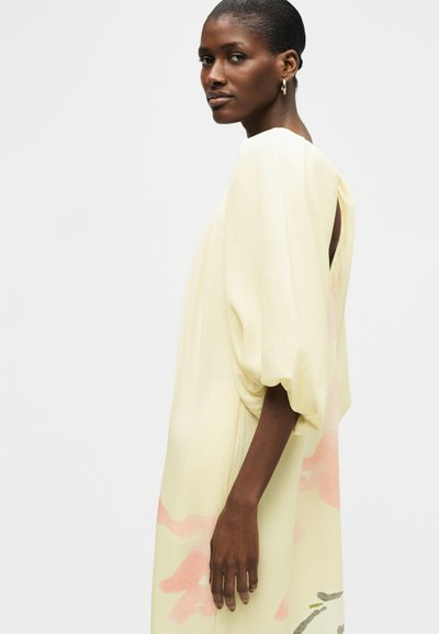 Γυναίκα με κοντά μαλλιά, φοράει ένα φαρδύ, ανοιχτό κίτρινο φόρεμα με ροζ και πράσινα αφηρημένα μοτίβα, στέκεται πλευρικά μπροστά σε απλό φόντο.