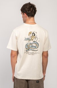 T-shirt en coton de couleur crème avec une impression graphique d'une femme portant un chapeau et un serpent, avec du texte en bas. Design à manches courtes.