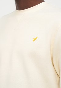 Κοντινό πλάνο ενός ατόμου που φοράει ανοιχτόχρωμο φούτερ με στρογγυλή λαιμόκοψη και μικρή κεντητή κίτρινη πουλίτσα στο στήθος.