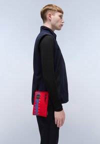 Gilet blu navy sopra una maglietta nera a maniche lunghe; il modello tiene una borsa a tracolla rossa con testo e zip blu. Tessuto liscio con un forte contrasto di colori.