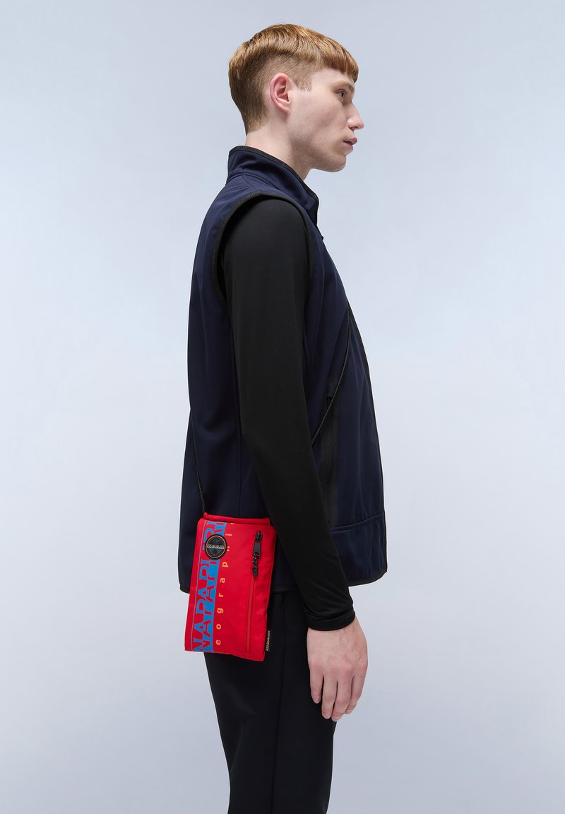 Gilet blu navy sopra una maglietta nera a maniche lunghe; il modello tiene una borsa a tracolla rossa con testo e zip blu. Tessuto liscio con un forte contrasto di colori.