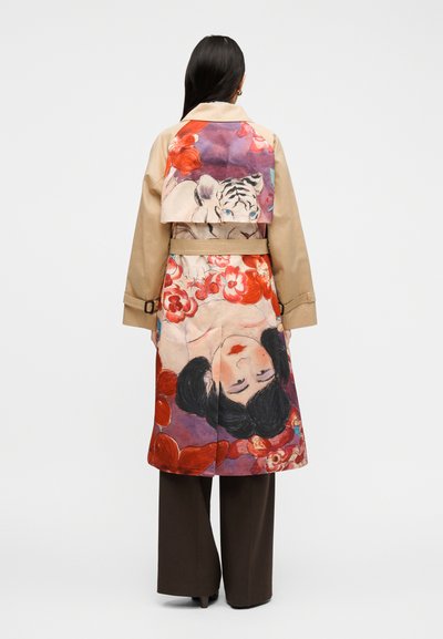 Femme portant un trench-coat beige avec un dos coloré présentant un visage de tigre blanc et un portrait de femme inversé entouré de fleurs rouges.