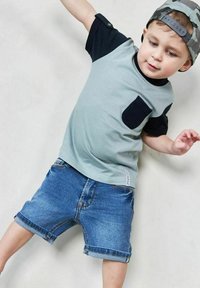 Vaaleanvihreä, lyhythihainen t-paita, jossa on tummansinisiä yksityiskohtia ja tasku, yhdistettynä sinisiin farkkushortseihin. Shortsit on käännetty lahkeista.