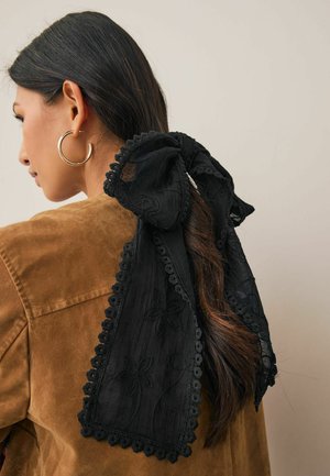 Frau mit langem, dunklem Haar, das mit einer großen schwarzen Spitzenbrosche zusammengebunden ist, trägt goldene Creolen und eine braune Wildlederjacke.