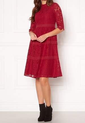 Femme portant une robe en dentelle rouge mi-longue avec des manches trois-quarts et des bottines noires, debout devant un mur blanc à panneaux.