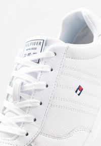 Weißer Sneaker aus glattem Leder mit tonalem Nähen, ausgestattet mit weißen Schnürsenkeln, einem gepolsterten Schaft und einem kleinen Emblem in Rot, Weiß und Blau.