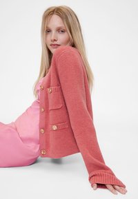 Rózsaszín kötött kardigán két elülső zsebbel, aranygombokkal és bordázott ujjvéggel. Egy rózsaszín szatén ruhadarab fölött viselve a kontraszt érdekében.